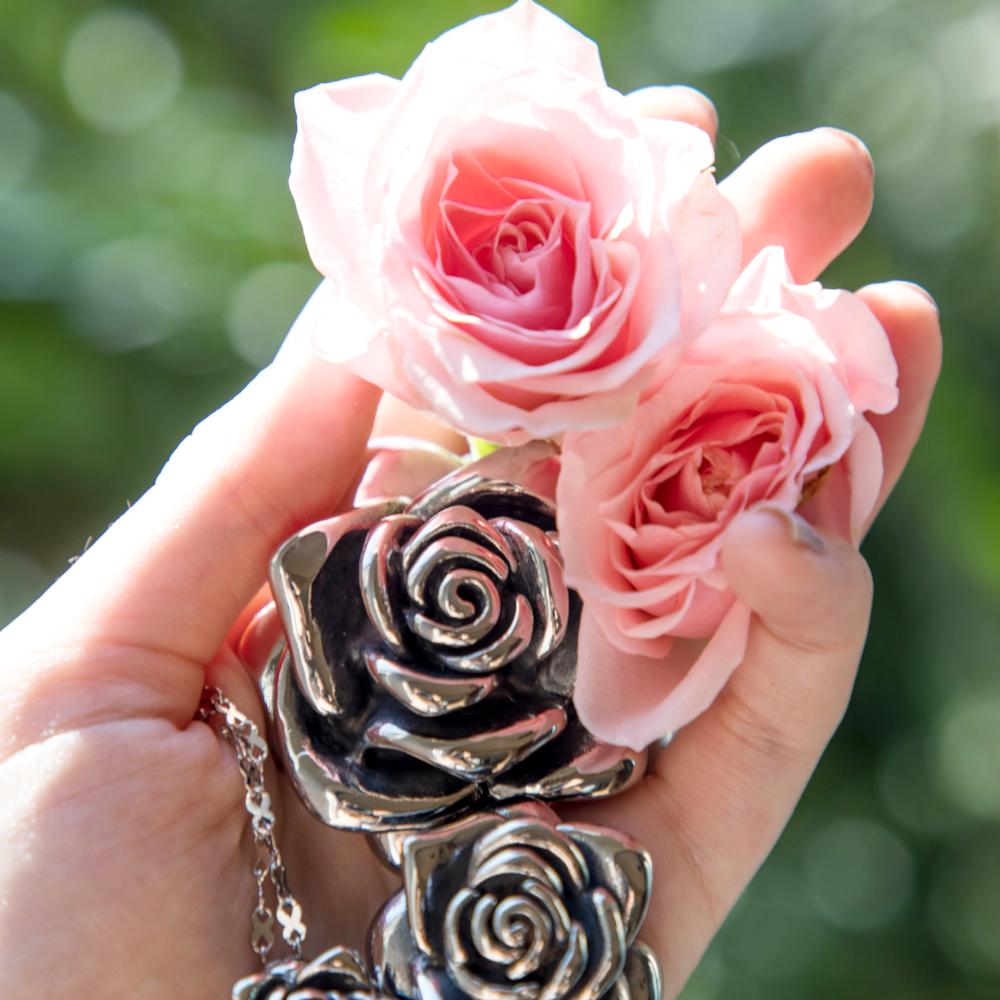 Large stainless steel rose pendant on our stainless steel infinity chain next to our medium pendant set by pink roses. This pendant will not tarnish or dent. Lovely rose flower pendant. The rose flowers are being held by a woman's hand.